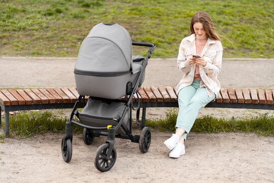 Positive, smiling mother using smartphone, sitting on bench, in stroller little baby. Woman walking