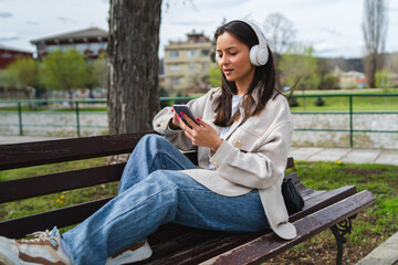 One young girl is listening to music on her wireless headphones and using her phone outdoors	
