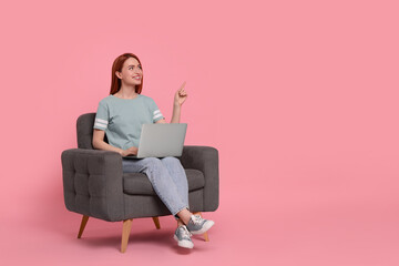 Fototapeta na wymiar Smiling young woman with laptop sitting in armchair on pink background, space for text