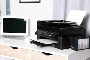 Modern printer with paper near laptop on white desk at home