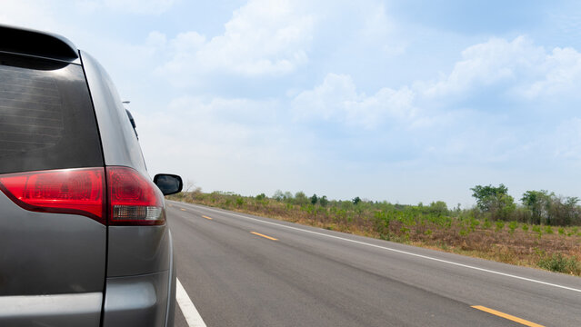 Rear Side Of Gray Car Triving On The Empty Asphalt Road. Beside With Wide Open Spaces Of Provincial Nature In Thailand. Under Blue Sky And White Clouds.