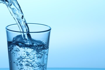 Pouring water into glass on light blue background, closeup. Space for text