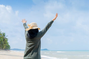 Happy Young muslim woman in grey dress and hat standing on the beach and open hands on sea with breaths a full breast and enjoys freedom.