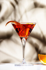 Martinez orange cocktail drink with red vermouth, liqueur, bitter, citrus zest and ice in martini glass. Light beige background, hard light, shadow pattern