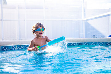 Cute African American little boy playing in a pool with a water gun launcher. Getting wet, spraying other people and Having lots of fun in the summertime outdoors