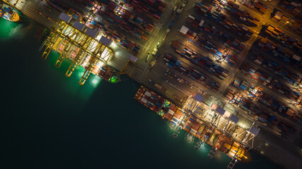 night scene commercial port loading and unloading cargo from container ship import and export by crane for distributing goods by trailers transported to customers and dealers, aerial top view