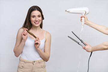 Smiling young woman holding hair styling tools, straightener and dryer, isolated on white background