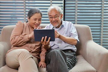 Happy smiling asian senior couple sitting on sofa and using tablet while online video call with friend or relative cousin at home living room. Internet information technology and lifestyle concept.