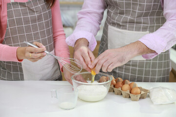 Happy young couple multiethnic man and woman hands making homemade bakery cake, pouring milk while breaking eggs and mixing flour into glass bowl in the kitchen. Family having fun while prepare food