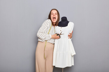 Happy positive female seamstress standing near mannequin with new dress and embracing it isolated over gray background, screaming with happiness, finishing outfit for new collection.