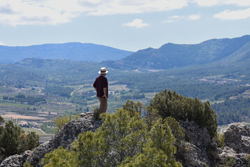 unrecognisable man in an explorer's cap, looking at the natural landscape on top of a small hill in a high place with a lot of countryside and forest.