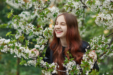Happy cheerful pretty smiling girl with red hair among blooming white flowers of cherry blossom and laughing in spring park or garden, outdoor lifestyle, tenderness, freshness, happiness of springtime