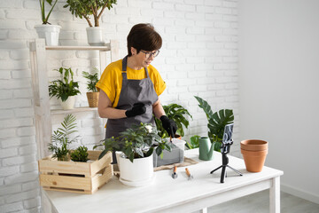 Relaxing home gardening. Smiling middle aged woman in black gloves with potted plant records gardening video blog in modern house - blogging and florist vlog influencer