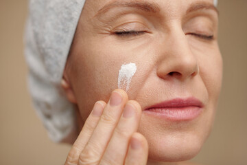 Mature woman applying moisturizing lotion on face after shower