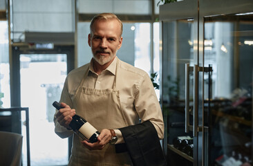 Portrait of mature waiter holding bottle of red wine and looking at camera while standing in restaurant