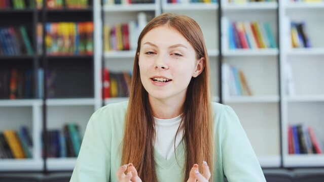 Caucasian girl talking and looking at camera in library. Female teenager recording vlog, streaming for social media, video calling in online chat. Web cam view headshot. Distance learning