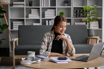 Happy Asian woman sits in her living room calculating taxes and managing household expenses.