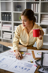 Focused Asian female accountant sipping coffee and analyzing business financial data