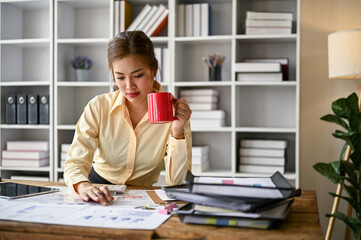Focused Asian female accountant sipping coffee and analyzing business financial data on reports