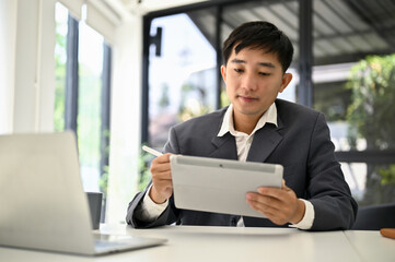 A focused Asian male boss using his tablet to manage his tasks at his office desk.