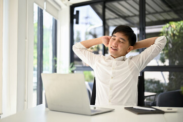 Tired Asian businessman sits at his desk, stretching his arms to relax after finishing work.