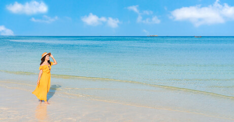 Beautiful young Asian woman wear a hat and yellow dress relax smile walking around sea beach with white cloud on blue sky in travel vacation. Summer and holiday concept. Free space