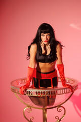 Pinup girl in a big glass like Dita Fon Tiz. Black wig, black underwear, stockings and red gloves, red light, red lipstick. Beautiful posch woman sitting in huge plastic goblet.