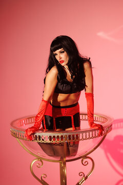 Pinup girl in a big glass like Dita Fon Tiz. Black wig, black underwear, stockings and red gloves, red light, red lipstick. Beautiful posch woman sitting in huge plastic goblet.