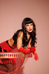 Pinup girl in a big glass like Dita Fon Tiz. Black wig, black underwear, stockings and red gloves, red light, red lipstick. Beautiful posch woman sitting in huge plastic goblet.