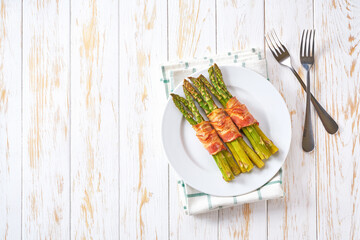 Baked  bundles of green asparagus wrapped in bacon in a ceramic plate on a white wooden table, top view.