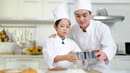 Asian father teaching little son preparing dough, baking cookies in kitchen wearing chef costume enjoying weekend playtime cooking baking. Happy family concept