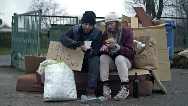 Two dirty and poorly dressed homeless people, a man and a woman, sit by a pile of rubbish with a handwritten LAST MY JOB HELP poster and eating something. Generative AI