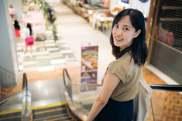 Smiling beautiful young asian woman looking at a camera while standing on an escalator in the shopping mall