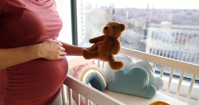 Pregnancy. Pregnant woman preparing nursery showing belly baby bump by crib holding teddy bear. Pregnancy concept and home nusery planning
