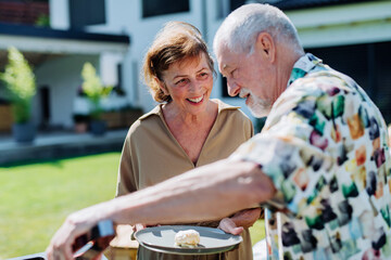 Senior man grilled outdoor at garden, giving his wife hamburger.