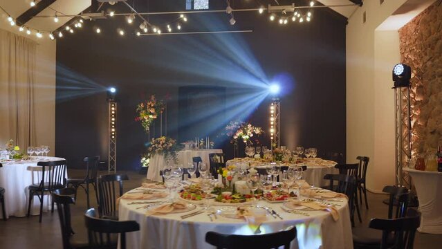 Festive banquet hall with decorated and served tables, working dynamic illumination from rotating beams and retro garlands. Rays hit lens, black chairs and wall, appliances are installed on farms.