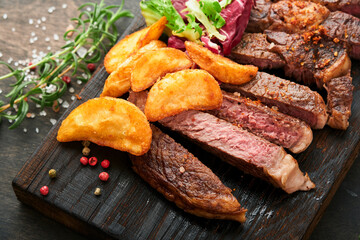 Steaks. Sliced grilled meat steak New York, Ribeye or Chuck roll with with garnished with salad and french fries on black marble board on old wooden background. Top view. Mock up.