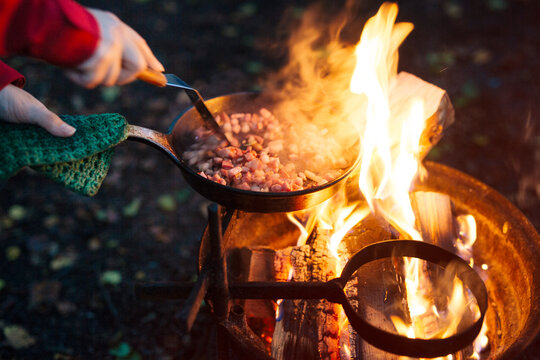 Person frying bacon in pan over campfire