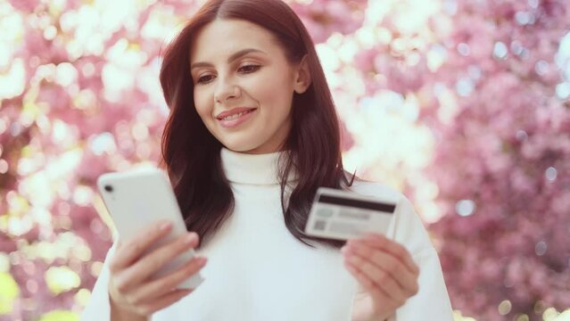 Portrait of young woman paying with credit card on smartphone with pink sakura blossom on the background Happy customer doing payments online shopping in internet store and receiving cashback outdoors