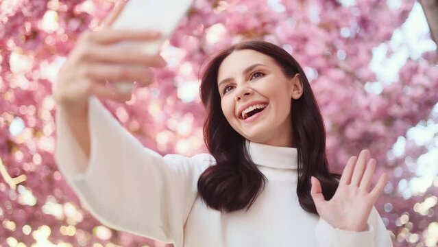 Portrait of smiling dark haired woman posing takes selfies and streams video to social media from her smartphone with pink sakura flowers blossom on the background outdoors