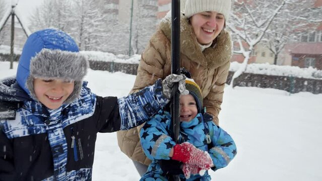 Happy cheerful mother with her two sons playing in snow at playground and riding on zipline. Fun and joy on winter holidays, family playing together, activity in snowfall