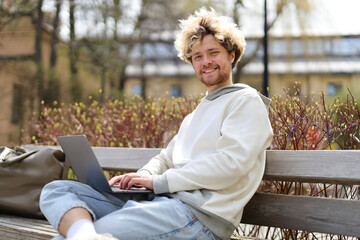 A smiling curly and bearded young man sits on a bench in the park and works on a laptop.