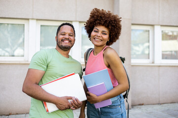 Two dark-skinned young millennials pose with textbooks and backpacks outside the university. The chubby Dominican man and the woman with the afro hair look at the camera happily. Multiethnic concept.