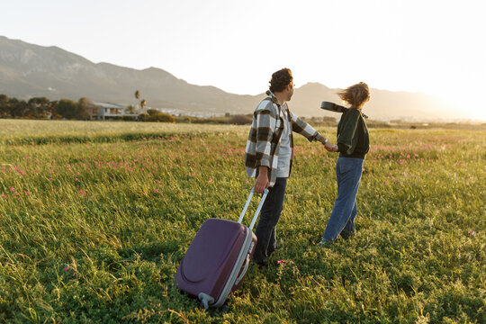 Inspired young loving couple is looking at living plan in future. They are standing with suitcase and ukulele and holding hands. Searching for new homeland