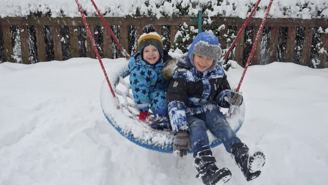 Happy smiling older boy with younger brother having fun on swing at public playground covered with snow after blizzard. Fun and joy on winter holidays, kids playing outdoors, activity in snowfall.