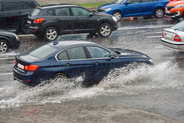 Fototapeta na wymiar Car splash water while driving on wet road, driving through puddle during heavy rain. Car driving on flooded asphalt road. Dangerous driving conditions, wet road risk of aquaplaning.