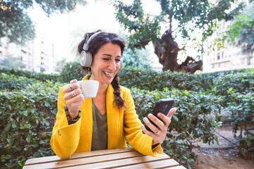 Smiling woman with wireless headphones with a coffee in her hand and looking at the phone