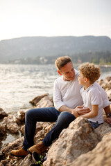 Brunette father and his blondie curly hair son sitting at looking to each other at the stones of Italian Garda lake 