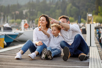 Cute stylish family sitting and smiling on the wooden pier and point with finger on Italian Garda...