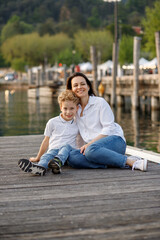 Brunette mom and blondie curly hair son sitting and smiling at the wooden pier in Italian Garda lake port 